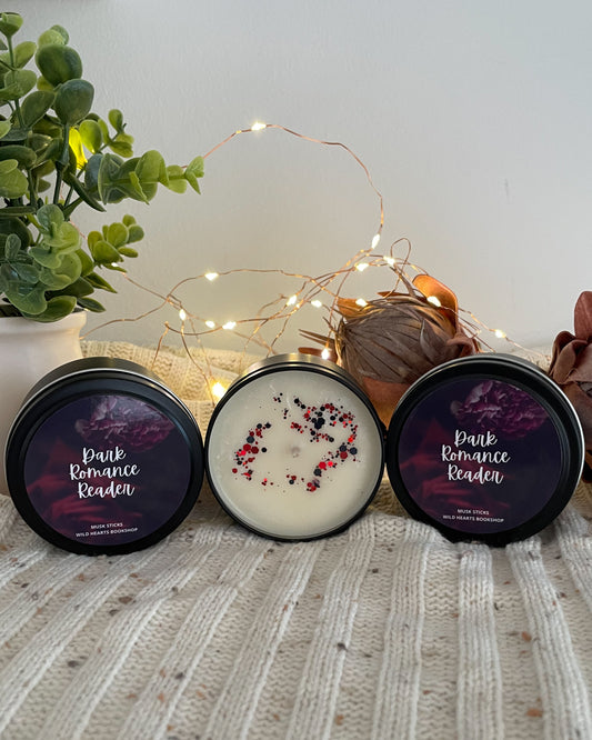 Three candles with decorative labels on a textured surface with a plant and lights in the background.